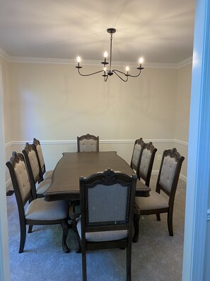 A dining room with a large wooden table surrounded by six upholstered chairs with dark wood frames and light fabric seats. A chandelier with multiple light bulbs hangs from the ceiling. - Dream House (Alexandria)