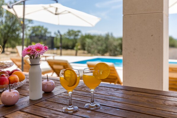 A wooden table with two glasses of amber-colored drink, a white vase with pink flowers, and a bowl of fruit.