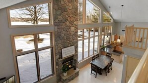 A spacious interior with large windows, a stone fireplace, and a kitchen area.