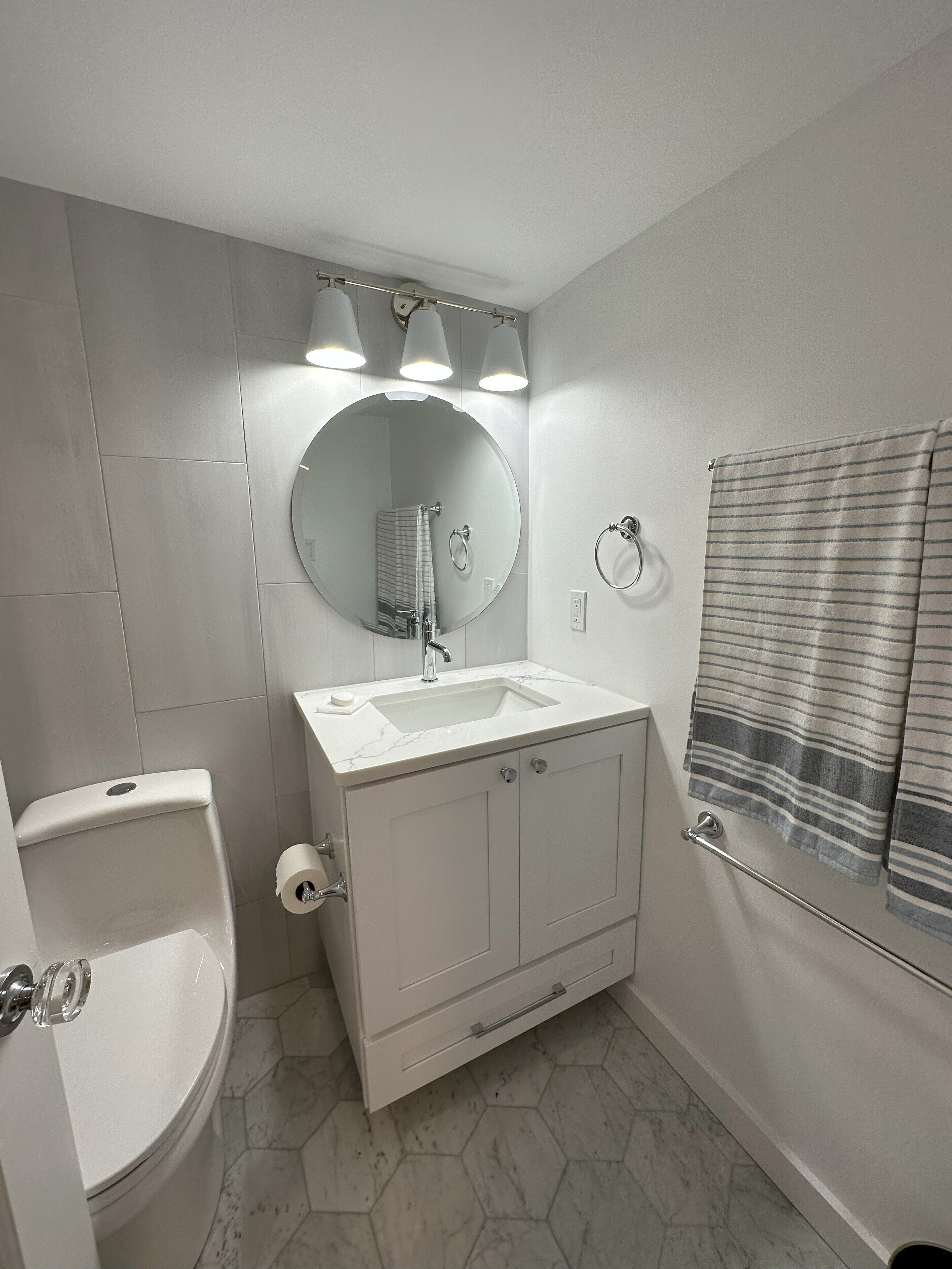 Een badkamer met een witte wasbak, spiegel en toilet. Drie lampen hangen boven de spiegel, en er is een handdoekrek met een gestreepte handdoek.