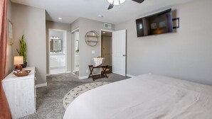 Une chambre à coucher comprenant un lit, un ventilateur de plafond, un téléviseur fixé au mur, un miroir et une porte donnant sur une salle de bain.