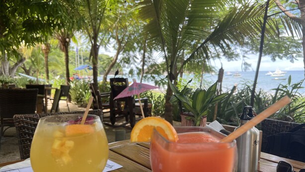 Deux boissons sur une table en bois, avec un menu à proximité, en plein air, avec vue sur la mer et les bateaux.