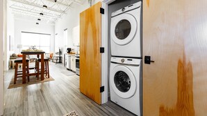 A laundry room with two stacked washing machines, a wooden door, and a kitchen area in the background. - The Cat's Meow:Midtown Bozeman (Bozeman)