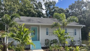 A single-story house with a blue door and windows, surrounded by various plants and trees.