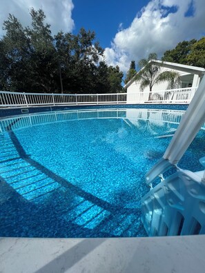 Une piscine d’un bleu éclatant, entourée d’une clôture blanche, bordée d’arbres et d’un bâtiment à la façade blanche.
