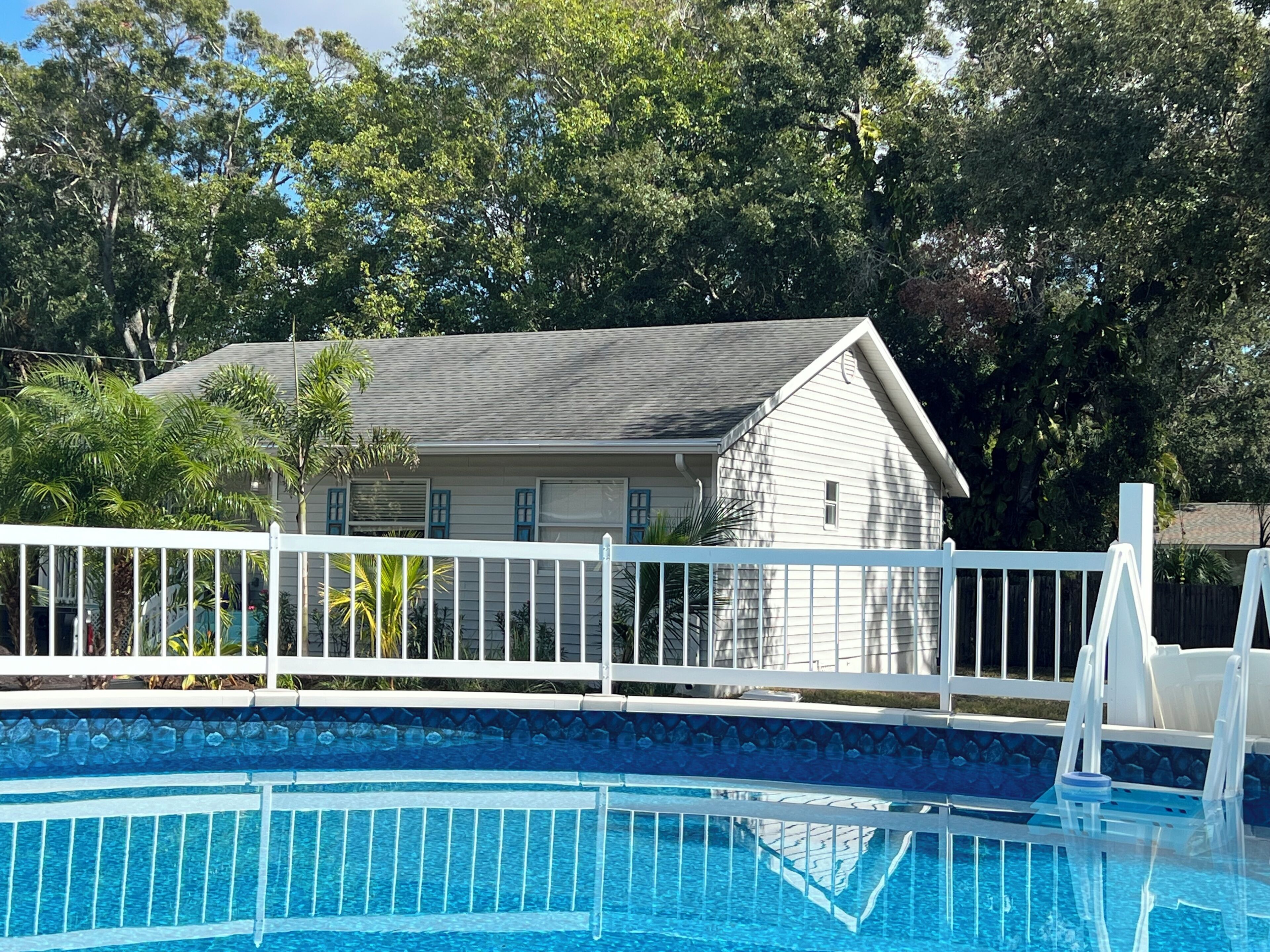 A swimming pool with a white fence, surrounded by trees and a house with a grey roof.