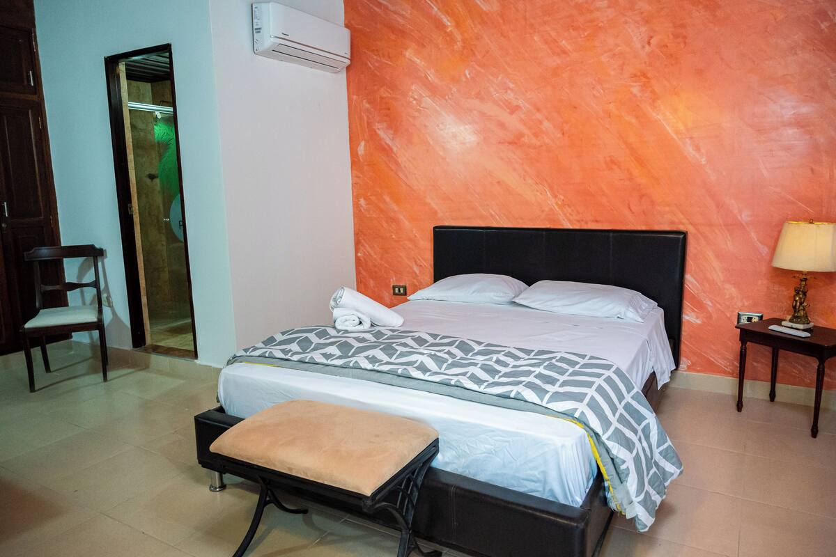 A hotel room with a bed, a chair, a table, and a wall-mounted air conditioner.