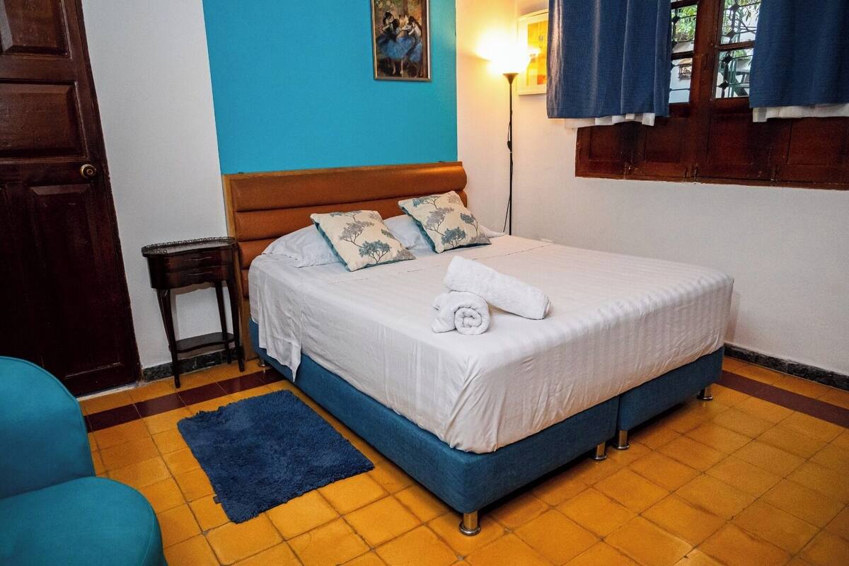 A bedroom with a bed, a nightstand, a blue chair, and a painting on the wall.
