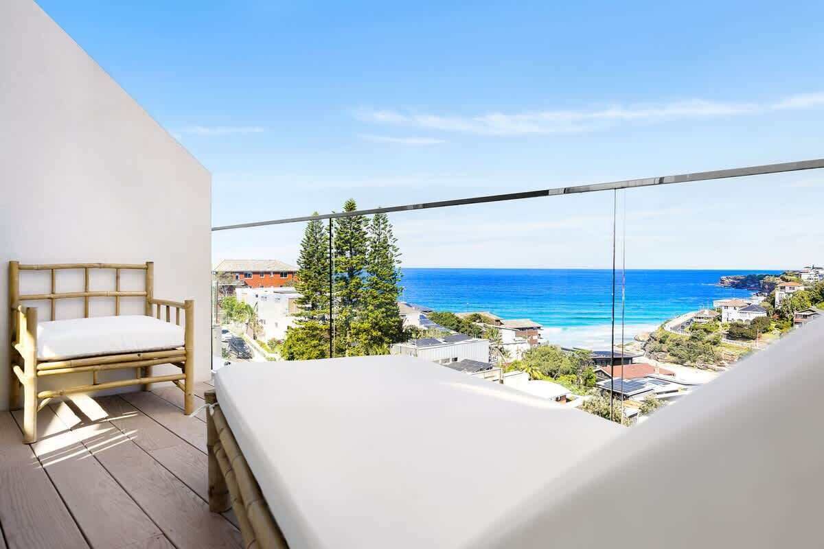 A balcony with a bed, a chair, and a view of the ocean and houses.