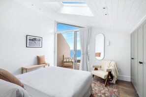 A bedroom with a bed, a chair, a bench, a mirror, and a view of the sea.