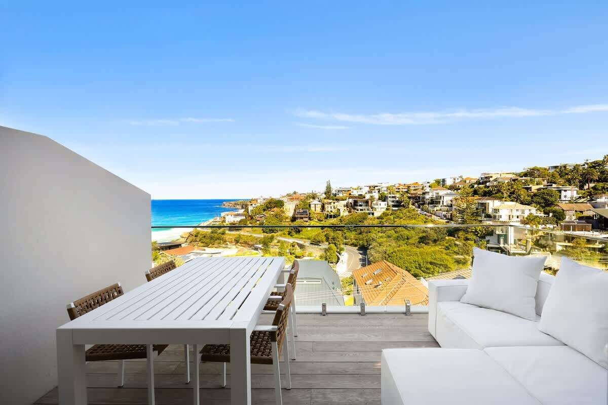 Un balcon avec une table et des chaises blanches, un canapé blanc et une vue sur une ville côtière.