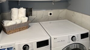 Un área de lavandería con dos lavadoras LG de carga frontal, una cesta de toallas dobladas y estantes con productos de limpieza.