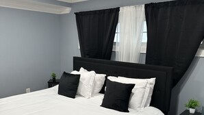 Un dormitorio con una cama grande, cortinas blanco y negro, una planta pequeña en la mesita de noche y un tapete oscuro.