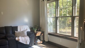 A living room with a dark gray sofa, a ceiling fan, a large window with curtains, and a wooden side table with a plant. - Burnash Bungalow - 4 min. to Fort Drum Gate (Felts Mills)