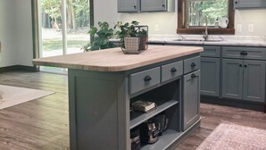 A kitchen with a central island, grey cabinets, a window with a view of greenery, and a built-in oven. - Burnash Bungalow - 4 min. to Fort Drum Gate (Felts Mills)