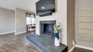 Una sala de estar con un televisor de pantalla plana sobre una chimenea, un comedor al fondo y un jarrón decorativo en la repisa.