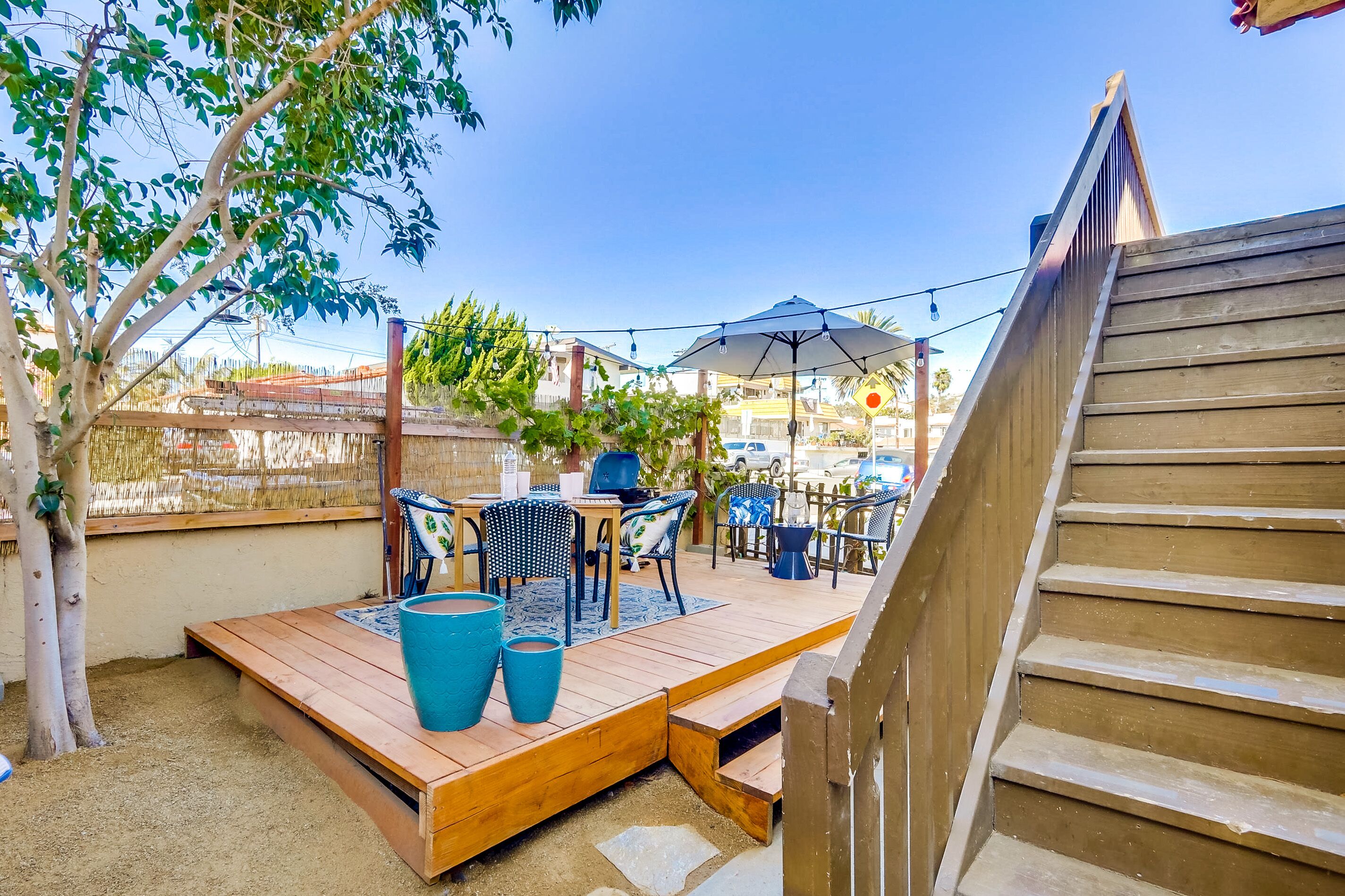Um deck de madeira com mobiliário para área externa, incluindo uma mesa e cadeiras, e um guarda-sol grande.
