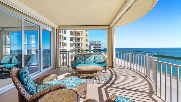 A balcony with wicker furniture and a glass door leading to a pool area. - Beach Colony Tower 14A | Oceanfront | Resort Amenities | Beach Getaways (Pensacola)