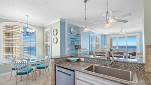 A kitchen with a granite countertop, stainless steel sink, and a dining area with a glass table and chairs. - Beach Colony Tower 14A | Oceanfront | Resort Amenities | Beach Getaways (Pensacola)
