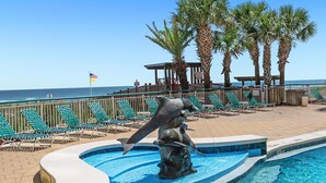 A pool area with a dolphin statue, lounge chairs, and a view of the ocean. - Beach Colony Tower 14A | Oceanfront | Resort Amenities | Beach Getaways (Pensacola)