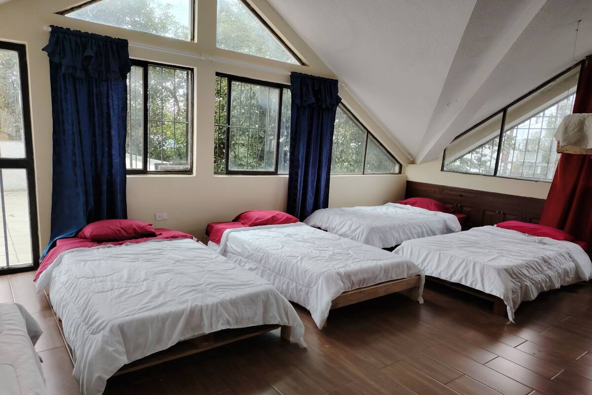 A room with four beds arranged in a row, large windows with curtains, and a sloped ceiling.