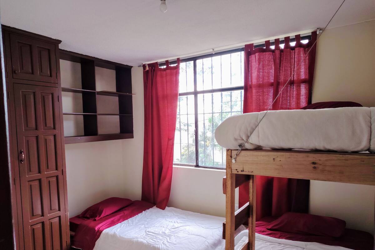 A dormitory room with a bunk bed, a wooden wardrobe, and a window with red curtains.