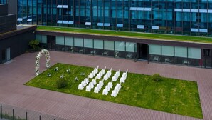 A modern building with a glass facade, a grassy courtyard with white chairs arranged in rows, and a decorative archway.