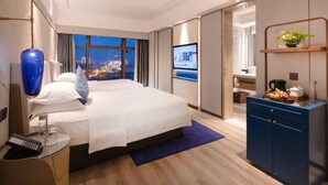 A modern hotel room with a large bed, a desk with a computer, a TV, and a view of the city at night.