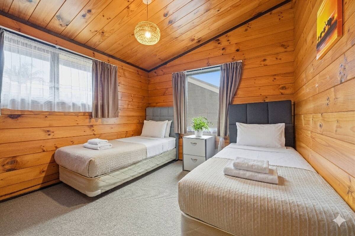 A wooden cabin room with two beds, a nightstand, a potted plant, and a ceiling light fixture.