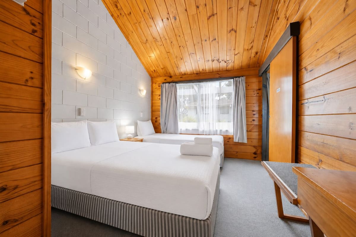 A room with a wooden ceiling, two beds, a window with curtains, and a table.
