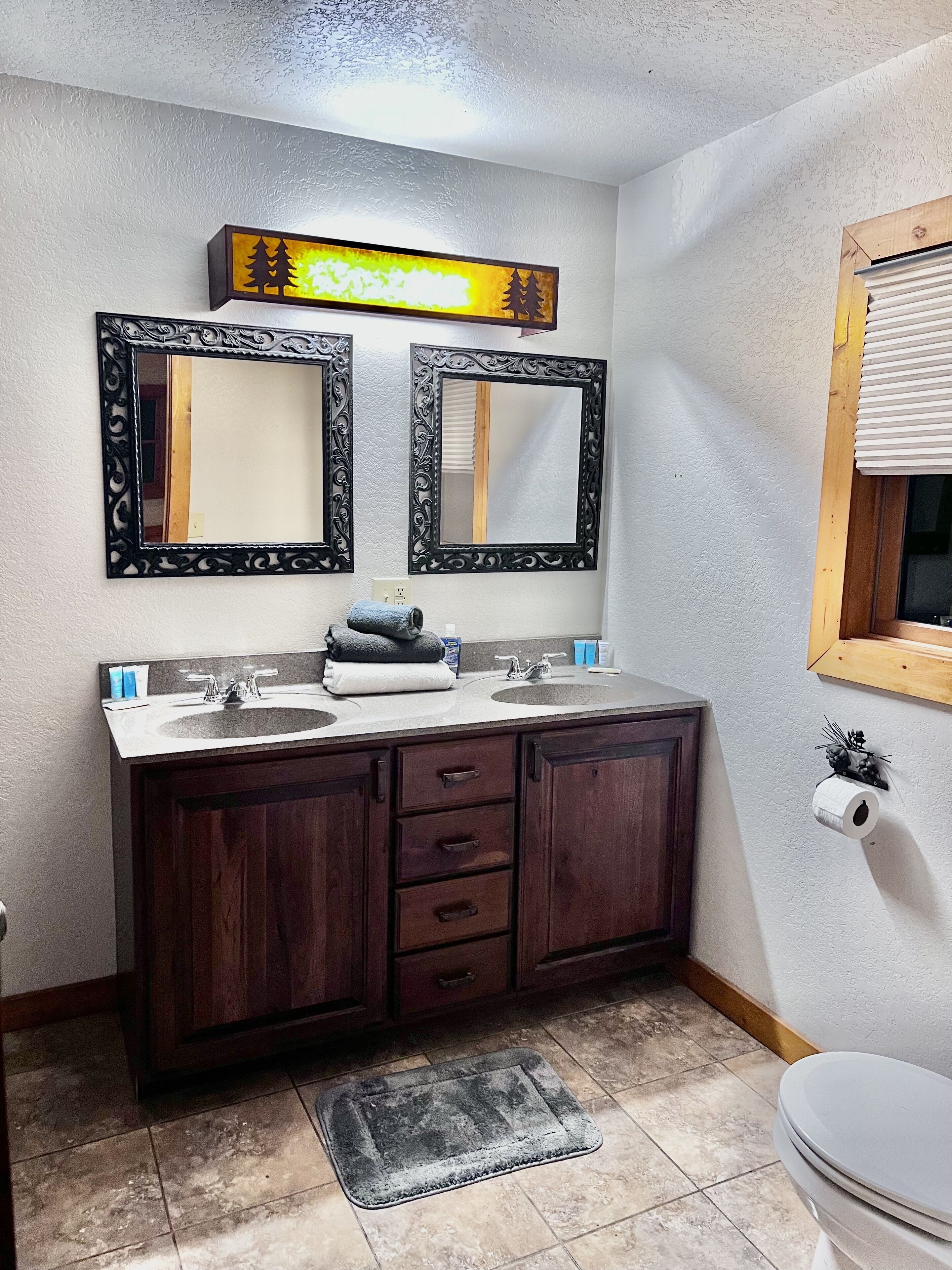 Un baño con dos lavamanos, muebles de madera oscura, un espejo con marco decorativo, un perchero para toallas y un inodoro con dispensador de papel higiénico.