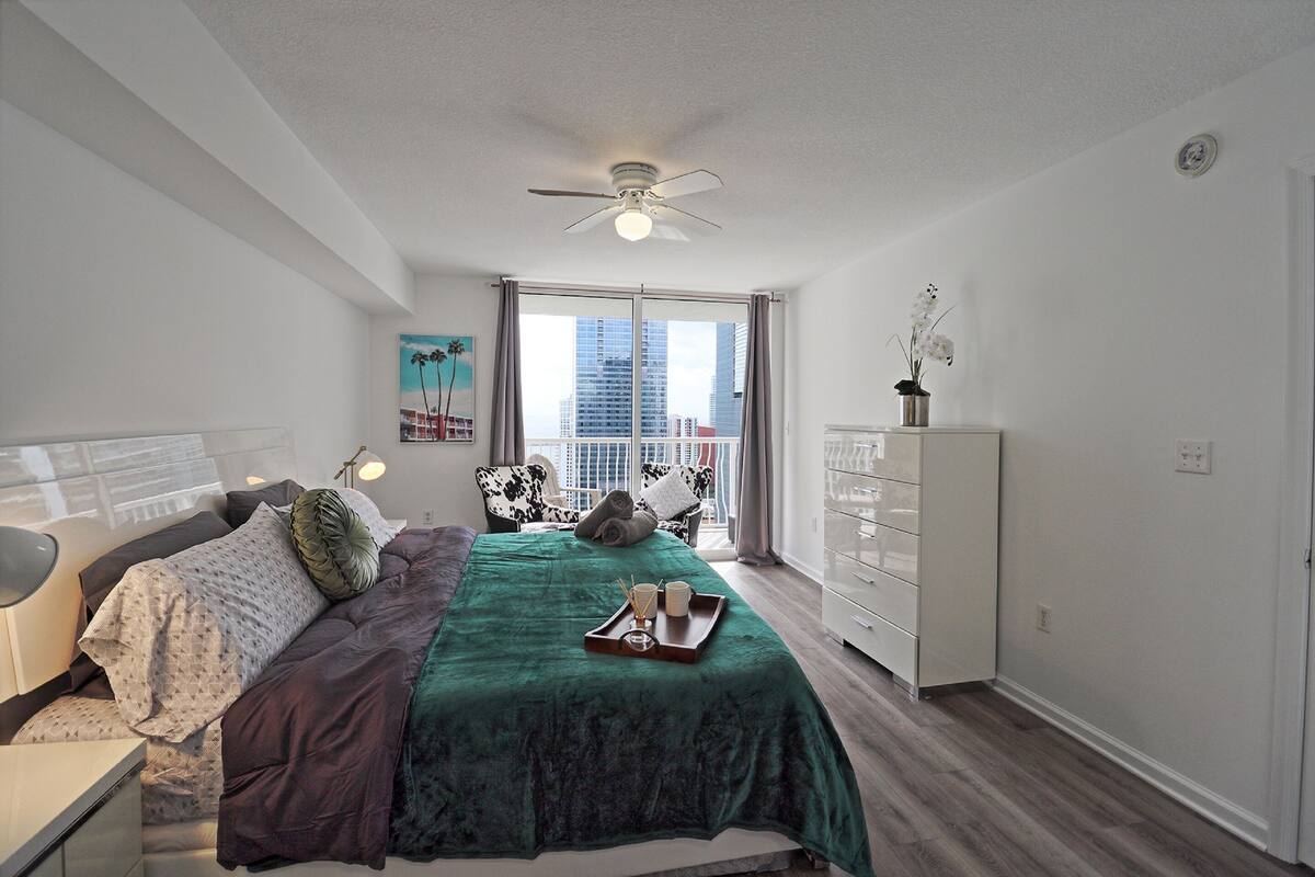 Quarto com cama, cómoda, ventoinha no teto e janela com vista para a cidade.