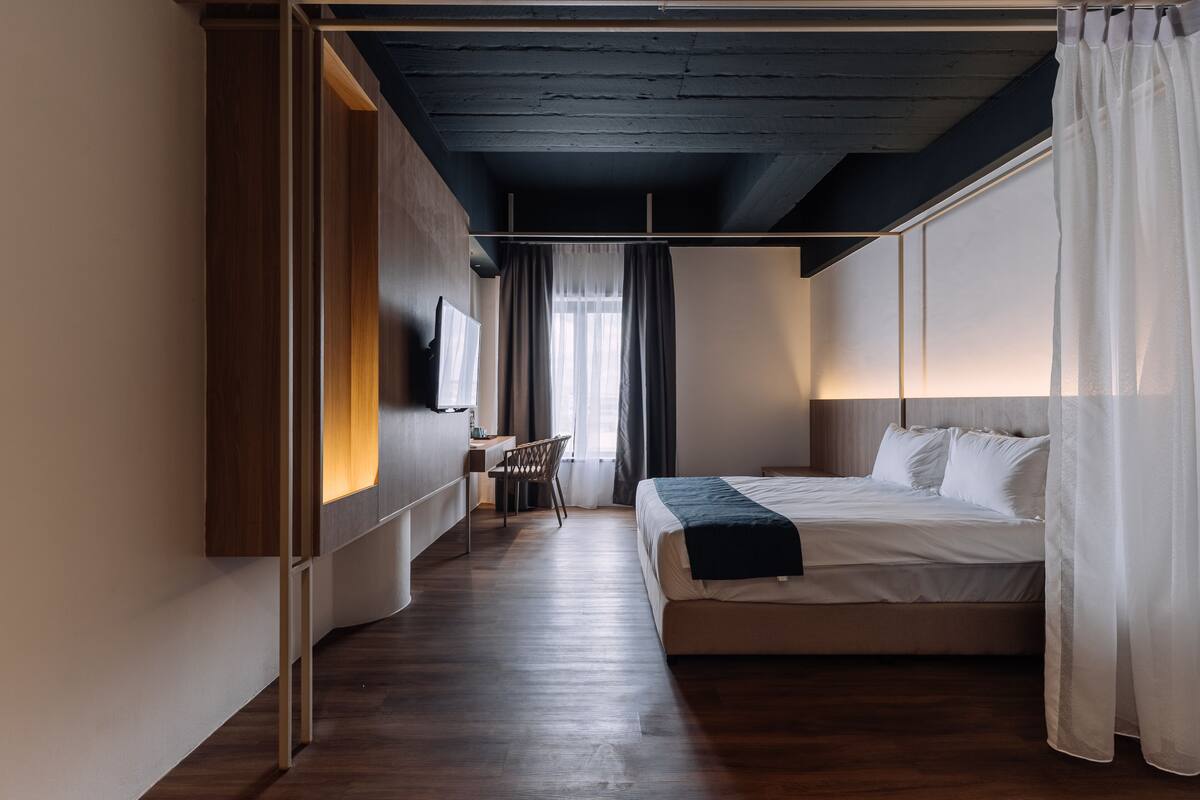 A modern hotel room with a wooden floor, a bed with white linens, a desk with a chair, and a TV mounted on the wall.