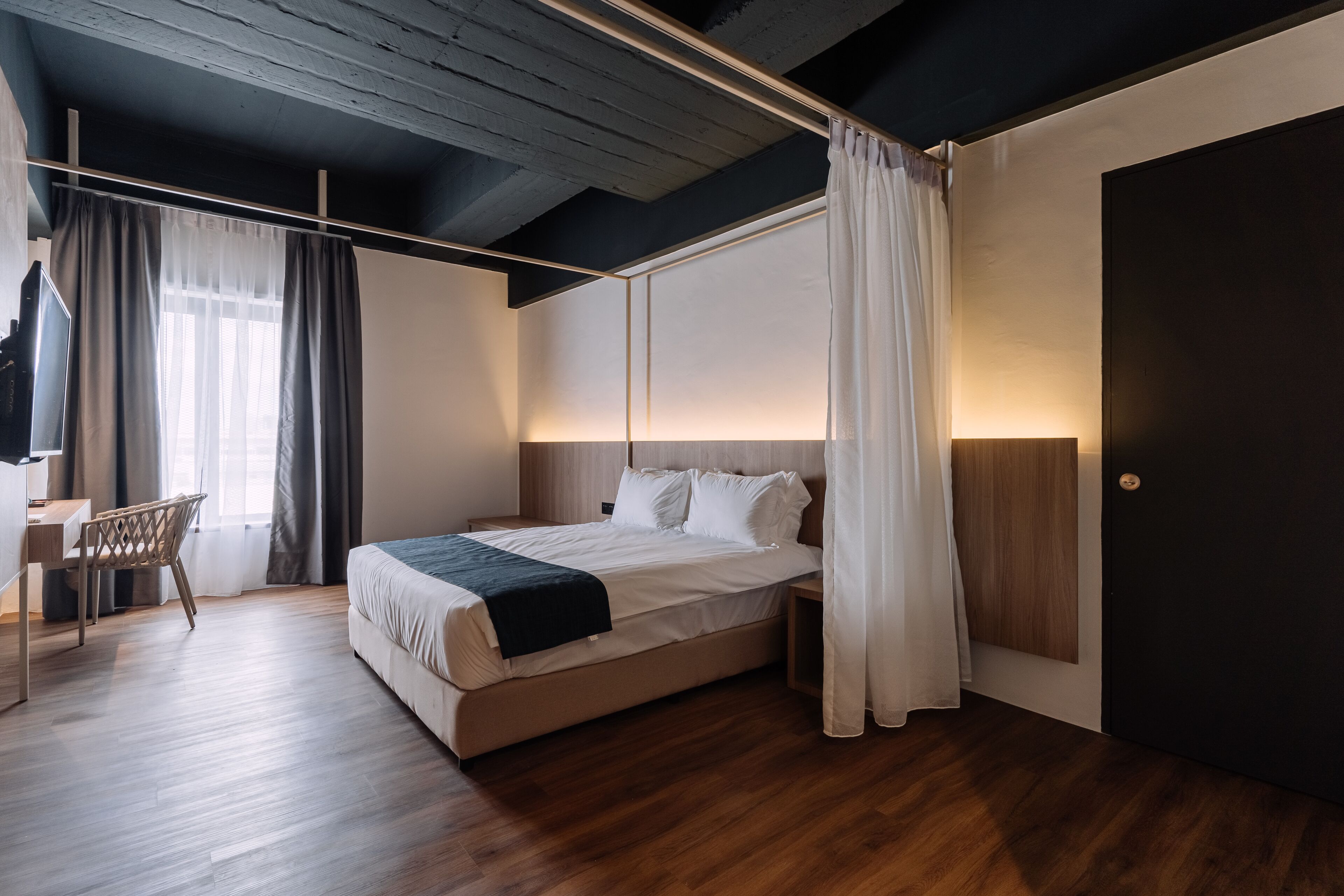 Quarto moderno com piso de madeira, cama com roupa de cama branca, escrivaninha com cadeira e uma janela grande com cortinas.