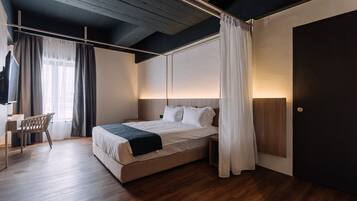 Quarto moderno com piso de madeira, cama com roupa de cama branca, escrivaninha com cadeira e uma janela grande com cortinas.
