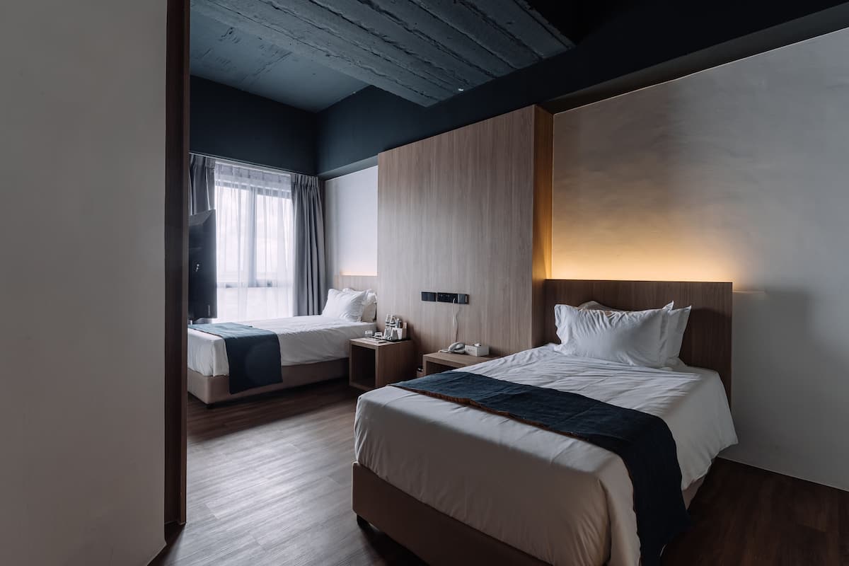 A hotel room with two beds, a wooden ceiling, a large window with curtains, and a bedside table with a lamp.