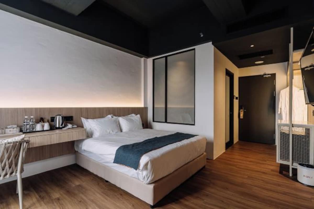 A modern hotel room with a large bed, a desk with a chair, a wardrobe, and a large window.