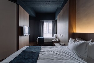 A hotel room with a bed, a TV, a bedside table, and a window with curtains.