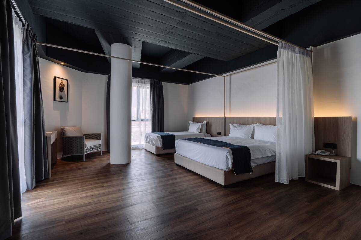 A modern hotel room with two beds, a small seating area, and a large window with curtains.