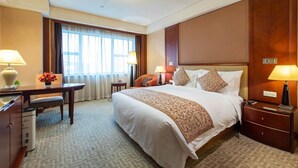 Quarto de hotel com uma cama grande, duas mesas de cabeceira, uma escrivaninha com cadeira, uma janela com cortinas e piso coberto por carpete.