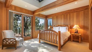A bedroom with a wooden bed, a chair, a nightstand, and large windows offering a view of trees.