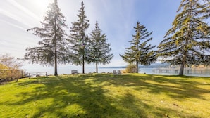 A grassy park area with benches and tall trees, overlooking a body of water and distant mountains.