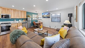A modern kitchen with wooden cabinets, a dining area with a table and chairs, and a living room with a sofa and cushions.