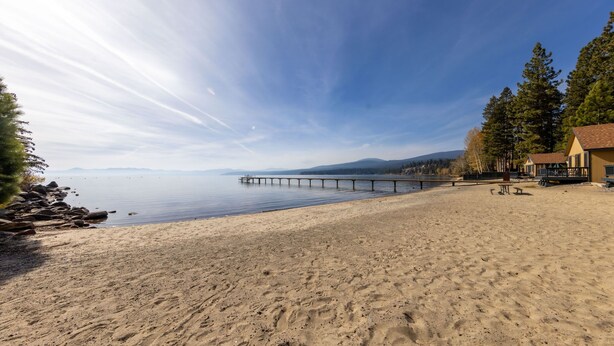 A beach with a wooden pier extending into calm waters, surrounded by trees and a building with a red roof.