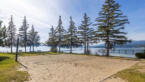 A sandy beach volleyball court with a net, surrounded by tall trees and a lake view.