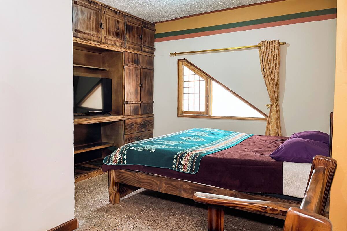 A bedroom with a wooden bed, a TV on a wooden cabinet, and a window with a curtain.