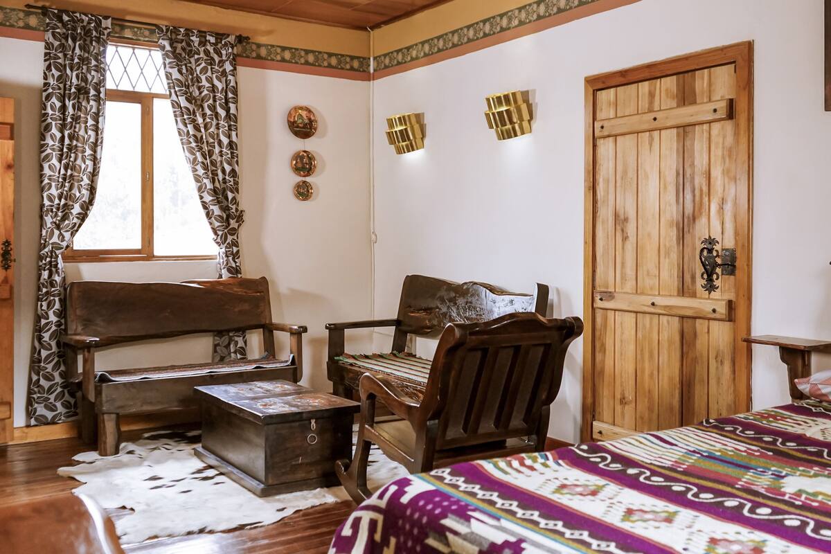 A cozy room with a wooden bed, a wooden chair, a small table, and a window with patterned curtains.