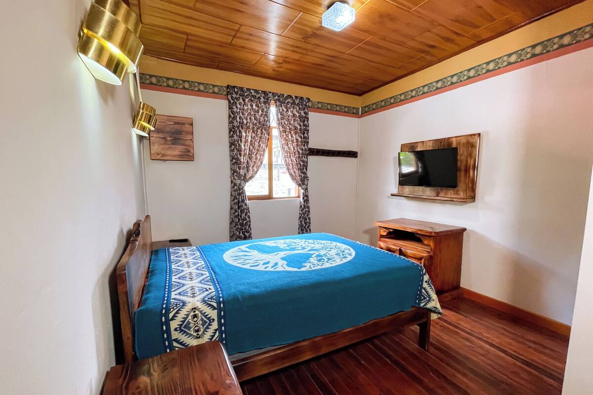 A bedroom with a wooden ceiling, a bed with a blue and white patterned bedspread, a wooden nightstand, a TV mounted on the wall, and a window with patterned curtains.