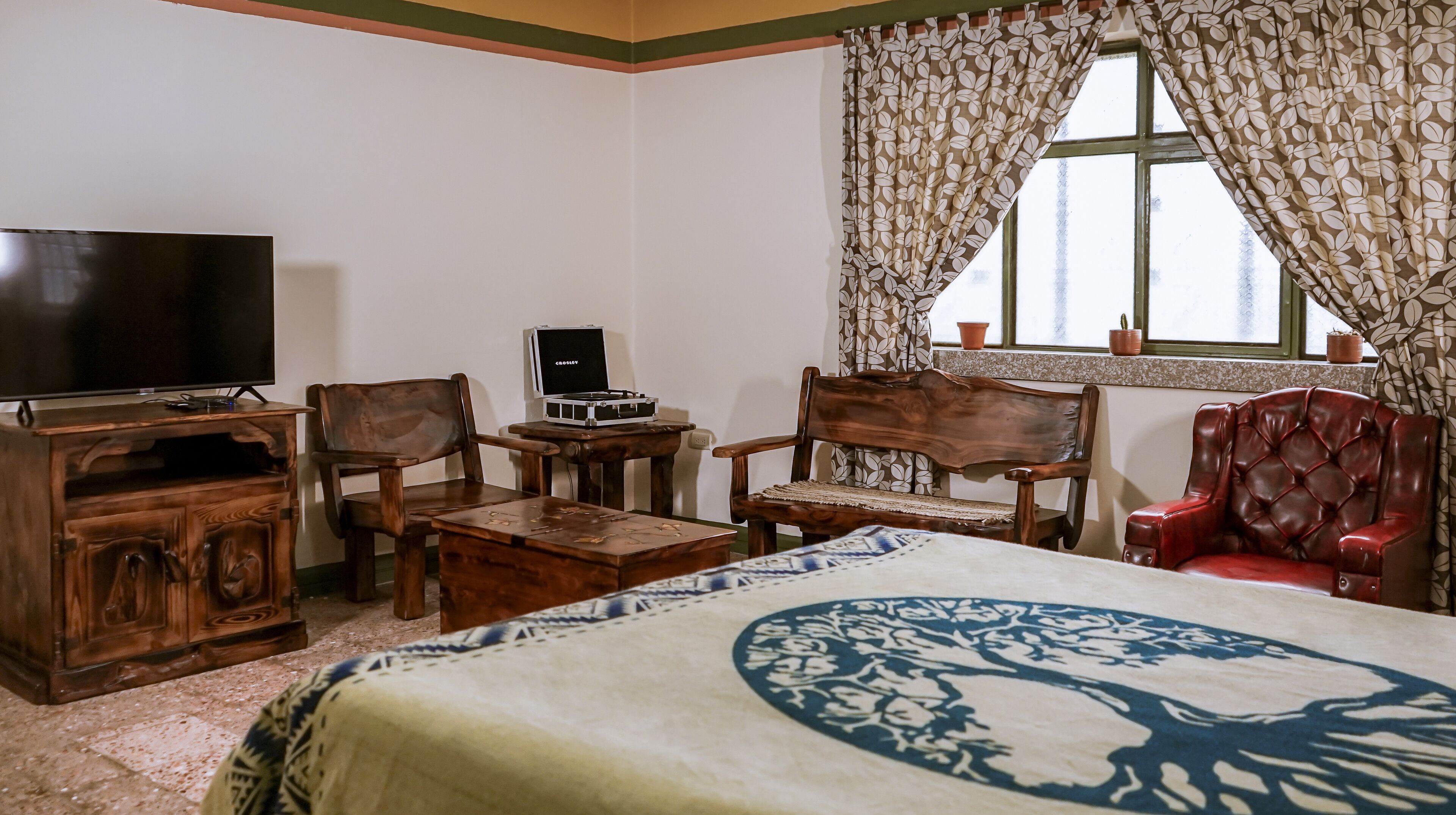 Quarto com cama, televisão, escrivaninha com computador, duas cadeiras de madeira e uma cadeira vermelha, além de janela com cortinas.