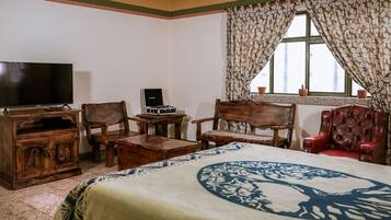 Quarto com cama, televisão, escrivaninha com computador, duas cadeiras de madeira e uma cadeira vermelha, além de janela com cortinas.